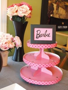 three tiered cake stand with pink flowers in vases behind it and barbie sign on top