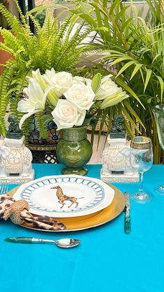 a table set with plates, silverware and vases filled with white flowers on a blue table cloth