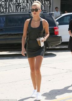 a woman walking down the street in a short black dress and white tennis shoes, wearing sunglasses