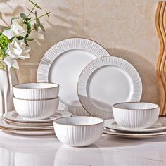 a white dinner set on a table with flowers in the vase and other dishes around it