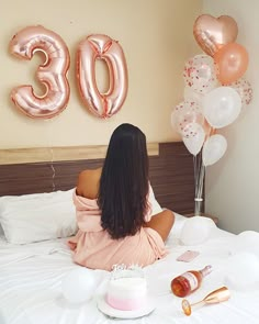 a woman is sitting on her bed with balloons and a cake in front of her