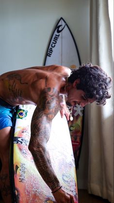 a man standing next to a surfboard with tattoos on his arm and chest, leaning over it