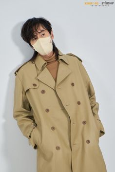 a man in a trench coat with a surgical mask on his face standing against a white wall