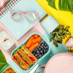 a lunch box with sandwiches, fruit and veggies next to a spiral notebook