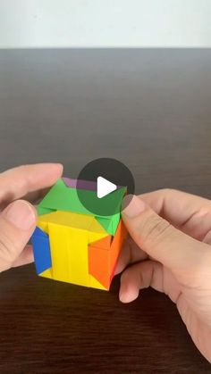 two hands are holding a colorful cube on a wooden table and one hand is pointing at it