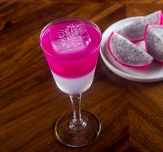 a pink drink in a glass next to some sliced fruit on a white plate and wooden table