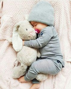 a baby sleeping with a stuffed animal on top of it's back and wearing a hat