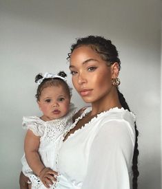 a woman holding a baby wearing a white dress and braids on her head, in front of a gray wall