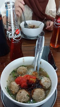 a bowl of soup with meatballs, tomatoes and broccoli next to two coke bottles