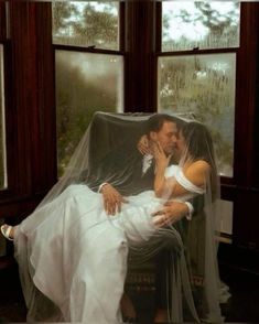 a bride and groom sitting on a chair in front of two windows, one kissing the other