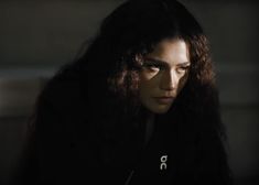 a woman with long dark hair stares into the distance while sitting in a dimly lit room