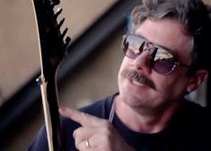 a man with sunglasses and a moustache points to something in his hand while holding an electric guitar