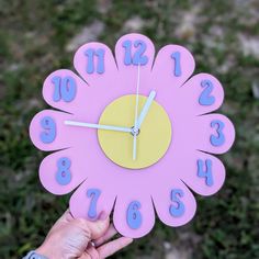 a hand holding a pink clock with blue numbers on it and a flower shaped face