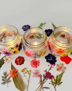 three glass jars filled with flowers on top of a white table next to leaves and candles