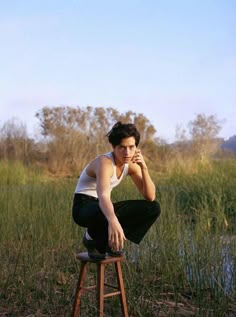 a young man sitting on top of a wooden stool next to a body of water