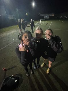 three girls posing for the camera on a soccer field at night with their hands up