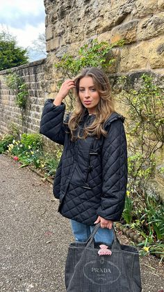 a young woman carrying a black bag on her shoulder while standing next to a stone wall