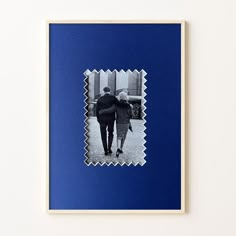 two people are walking down the street in front of a blue wall with a white frame