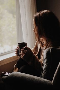 a woman sitting on a couch holding a coffee cup in her hand and looking out the window