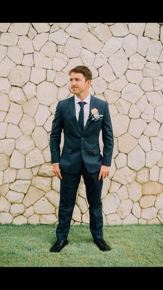 a man in a suit and tie standing next to a stone wall with his hands in his pockets