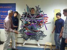 a group of people standing around a sculpture made out of strips of duct tape on a wall