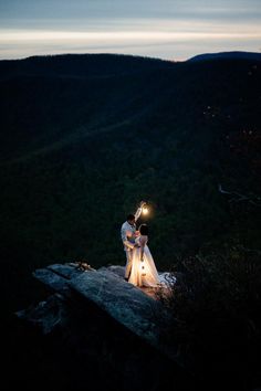 a bride and groom holding sparklers on top of a hill at night with mountains in the background