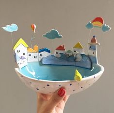 a hand holding a small bowl with houses and balloons floating in the sky above it