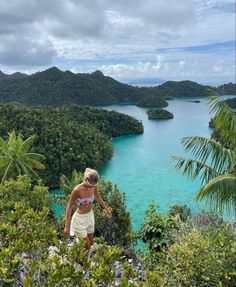 a woman standing on top of a lush green hillside next to trees and blue water