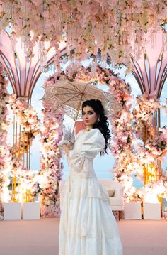 a woman in a long white dress holding an umbrella and posing for the camera with flowers all around her