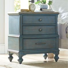 a night stand with two drawers and a clock on it's top, in front of a bed