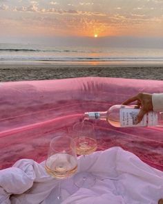 a person pouring wine into two glasses on a pink couch near the beach at sunset