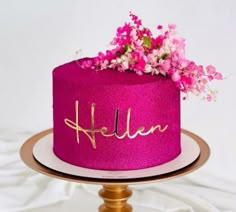 a pink cake with gold lettering and flowers on top is sitting on a wooden stand