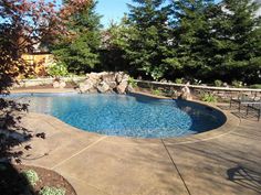 an outdoor swimming pool surrounded by trees and landscaping area with patio furniture, table and chairs