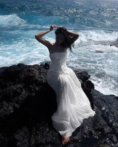 a woman in a white dress sitting on rocks by the ocean with her hands behind her head