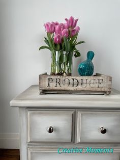 some pink tulips are in a vase on top of a white dresser with a sign that says produce