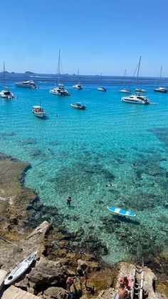 people are swimming in the clear blue water near boats on the ocean's shore