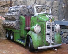 a green truck with logs on the back parked in front of a stone wall and tree stumps