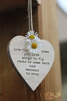 a white heart hanging from a wooden post with a daisy on it and the words give the love ones wings to fly roots to one back and reason to stay