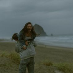 a woman carrying a child on her back while walking along the beach in front of an overcast sky