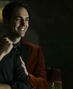 a man in a red jacket and black shirt smiles at the camera with his hand on his chest