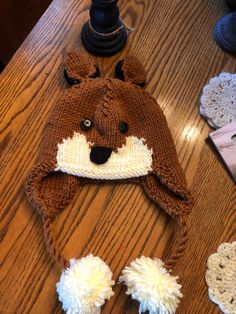 a brown and white knitted animal hat sitting on top of a wooden table next to doily