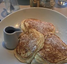 three pancakes on a white plate with syrup and sugar in the middle, next to a cup of coffee