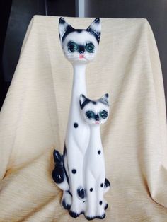 two ceramic cats sitting next to each other on a cloth covered table with a white sheet