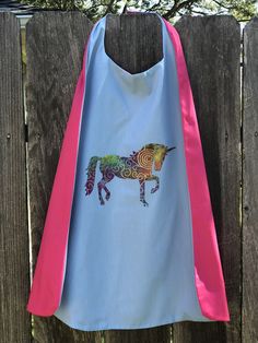 a blue and pink cape with a horse on it hanging from a wooden fence next to a tree