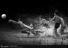 three boys playing in the water with a soccer ball and splashing water on them