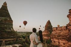 a man and woman standing next to each other near hot air balloons in the sky