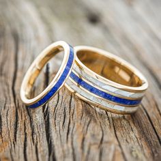 two gold wedding rings with blue and white stripes on them sitting on a piece of wood