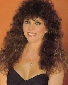 a woman with long curly hair wearing a black dress and smiling at the camera while standing in front of an orange wall