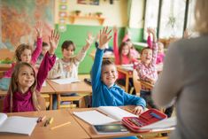 children raising their hands in a classroom with desks full of books and pencils