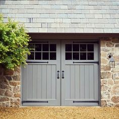 two gray double doors in front of a stone building with trees and bushes on either side